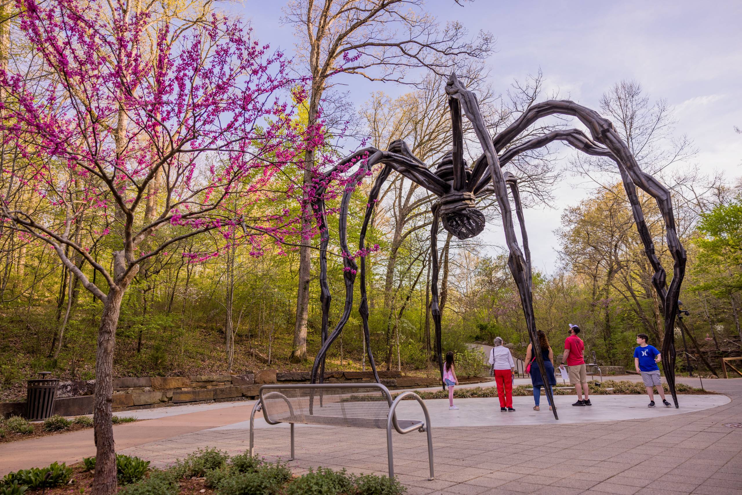 Maman (1) Large spider sculpture in park with people, trees, pink flowers, and a nearby bench.