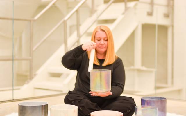 Woman seated on floor playing colorful crystal singing bowls with a mallet in an indoor setting.