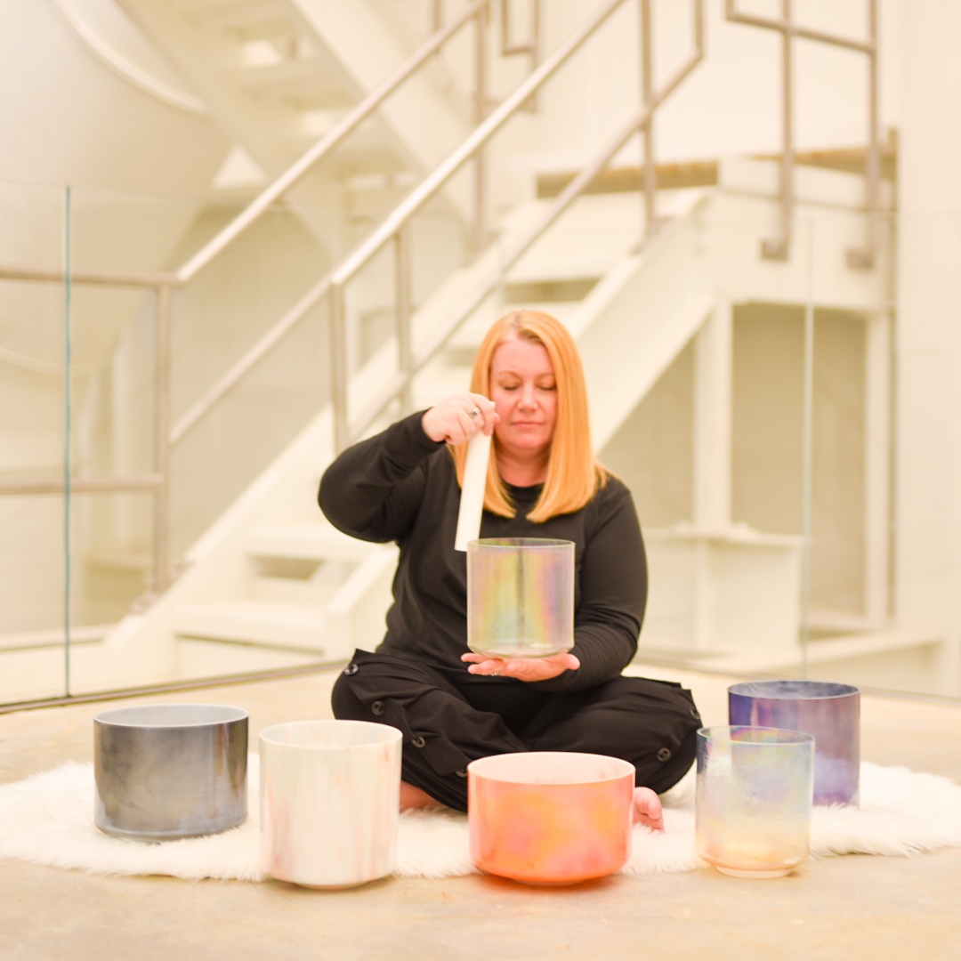 Meditate Woman seated on floor playing colorful crystal singing bowls with a mallet in an indoor setting.