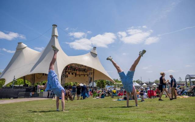 People performing handstands on grass at an outdoor event with tented stage and attendees nearby.
