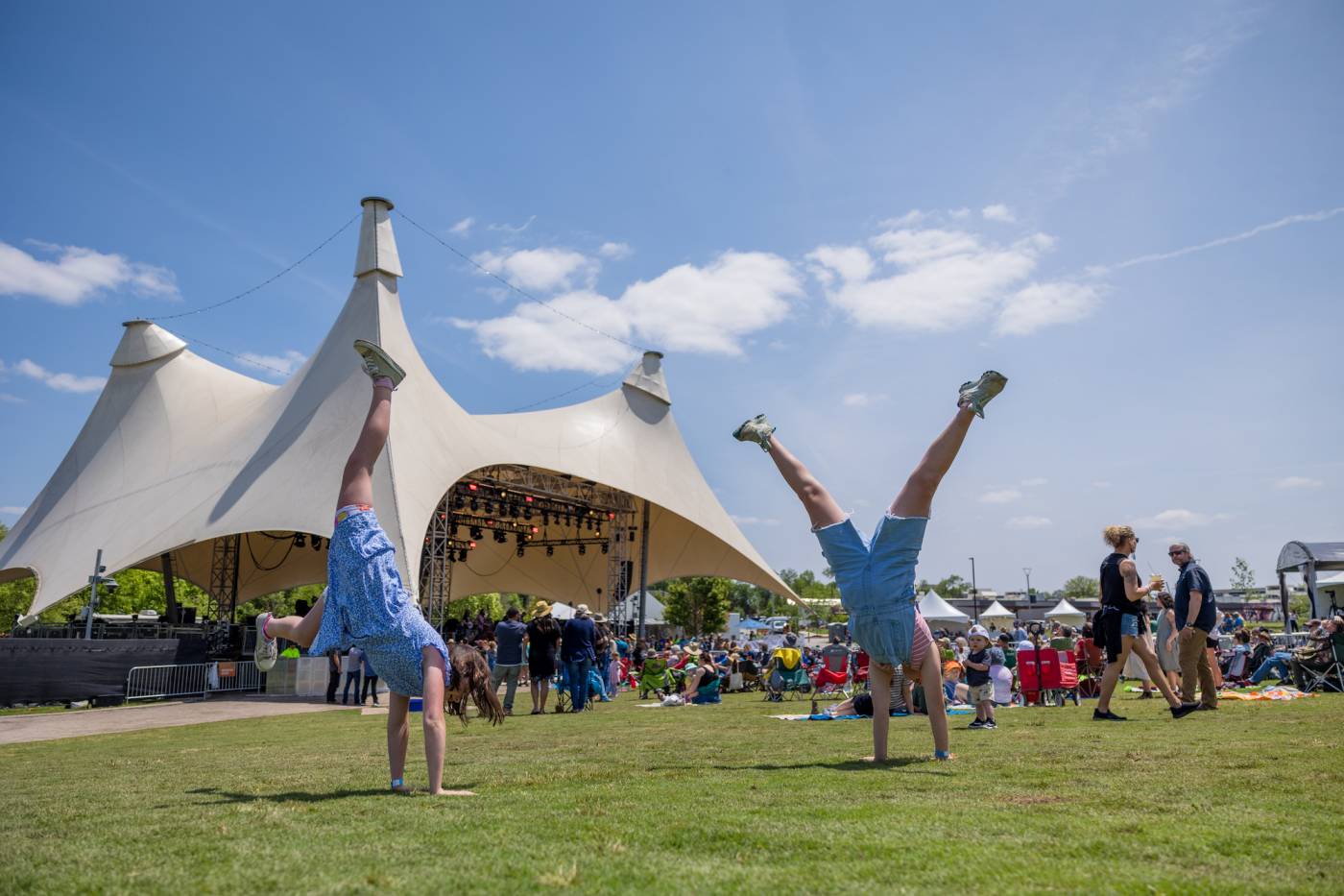 Mo green People performing handstands on grass at an outdoor event with tented stage and attendees nearby.