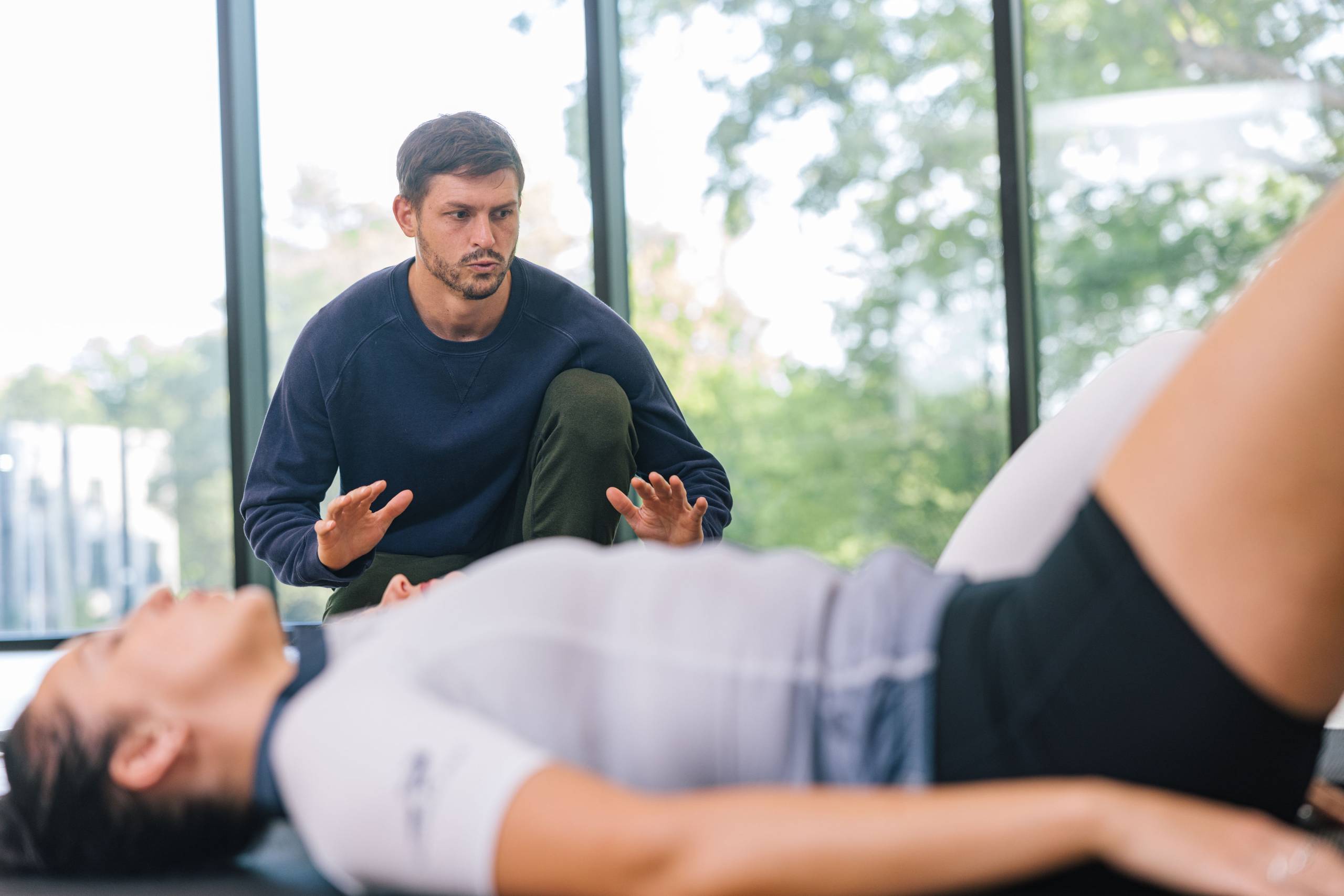 Mopros Person in blue sweatshirt kneels, guides another lying down in bright room with large windows.
