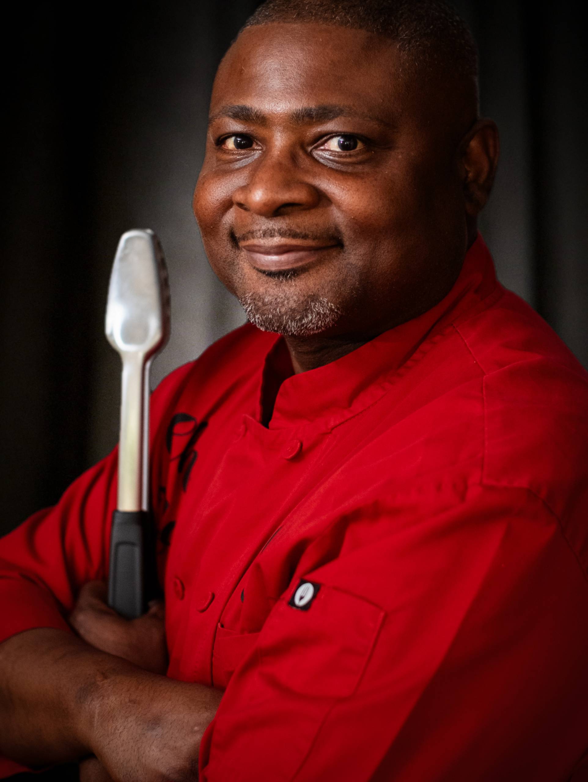Chef Nate Walls Person in red chef's jacket holding metal kitchen tool, dark background.