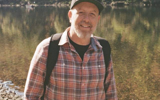 Man in plaid shirt and cap by a lake, forest background, wearing a backpack, smiling.
