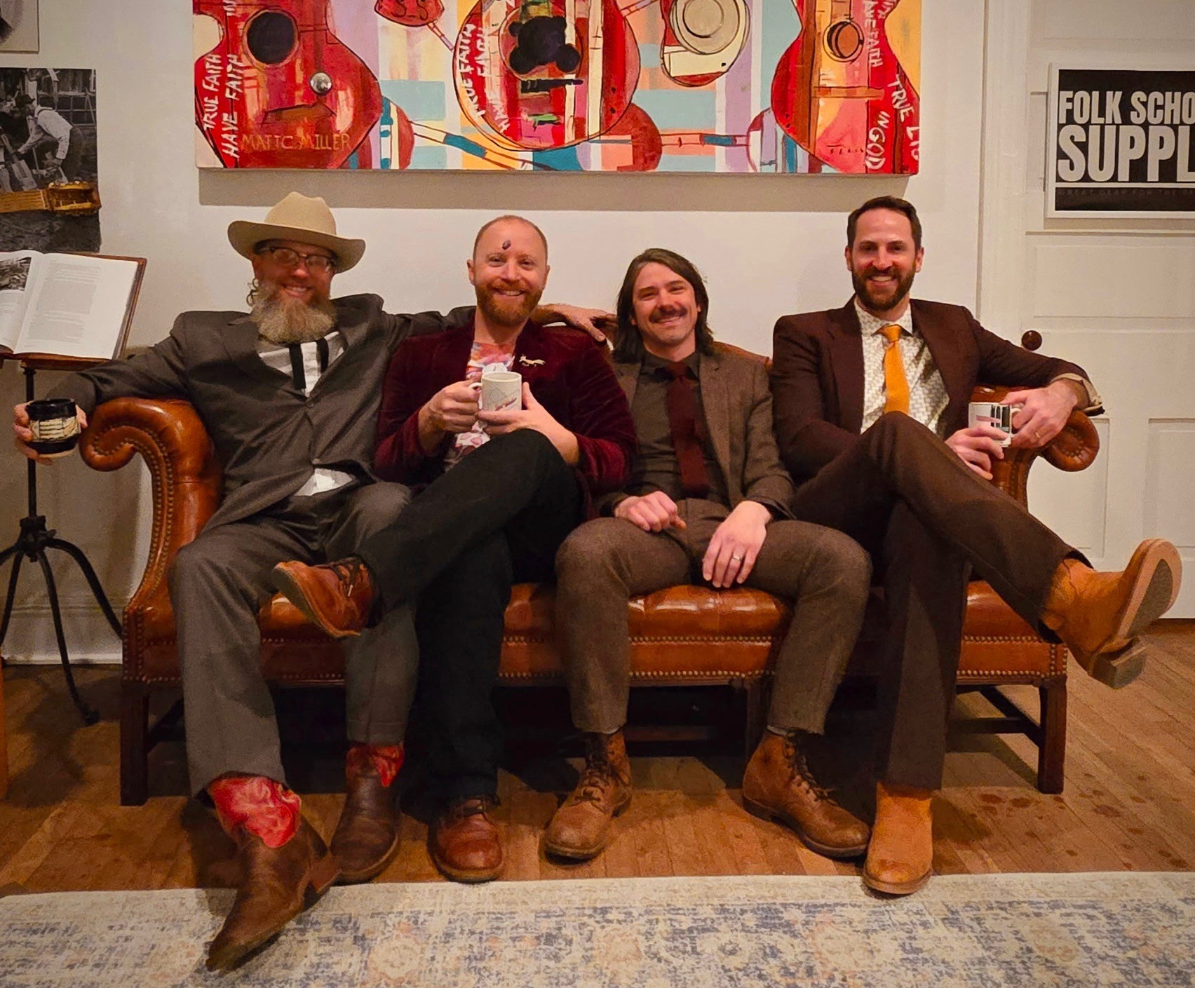 Four men in suits sit on a brown leather couch indoors holding mugs with artwork behind.