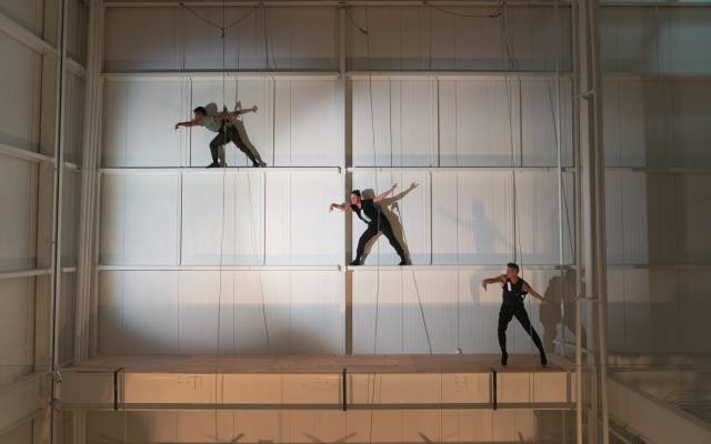 Performers suspended on wall with harnesses in well-lit indoor space, creating dynamic poses.