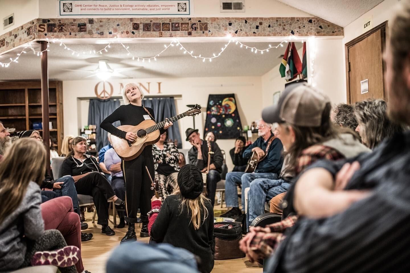 The Listening Circle Person playing guitar in a cozy room with seated audience and string lights.