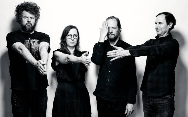 Four individuals in dark clothing pose with varied hand gestures against a white background.
