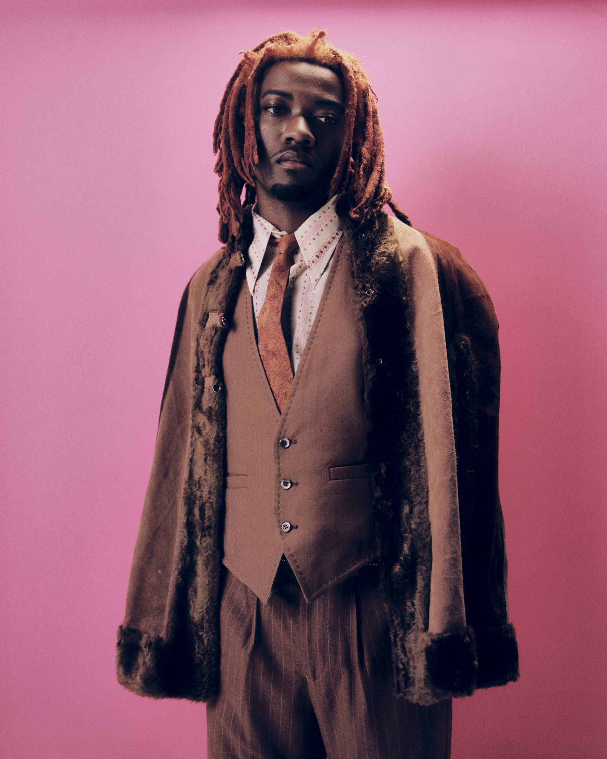 Man with reddish dreadlocks wearing a brown fur-lined coat, vest, tie, and pinstriped pants against pink background