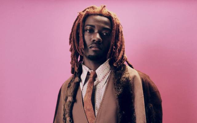 Man with reddish dreadlocks wearing brown layered coat, vest, tie, and pinstriped pants against pink background