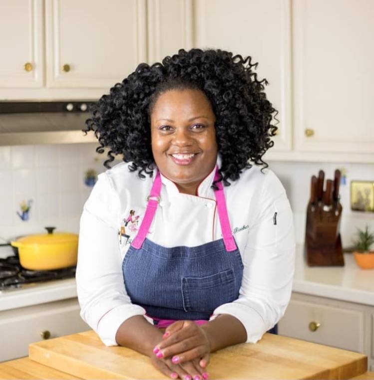 Chef Jennifer Hill Booker Chef Jennifer Hill Booker in a white coat and pink apron in a kitchen with white cabinets and a yellow pot on stove.