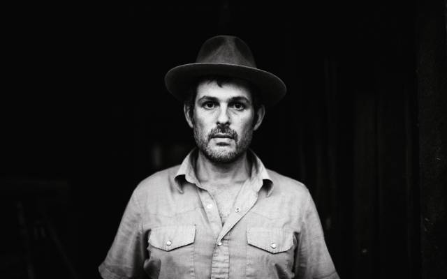 Black and white portrait of a person in a hat, button-up shirt, dark background.