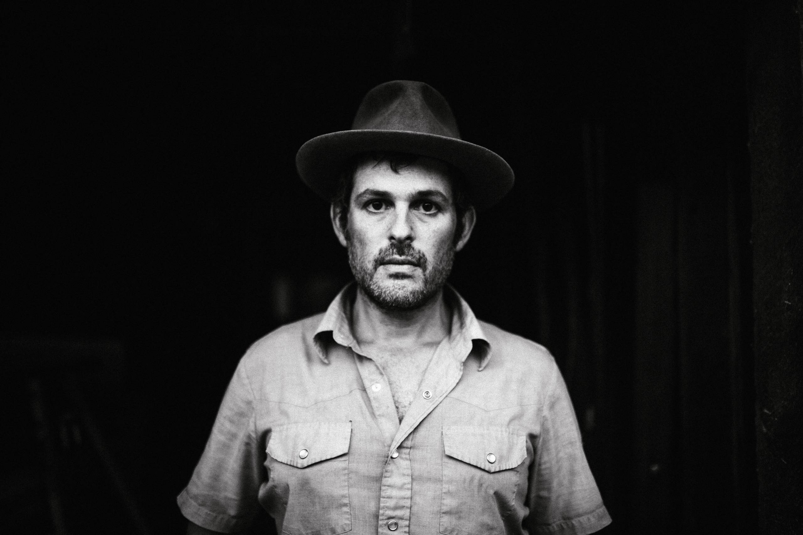 Gregory Alan Isakov Black and white portrait of a person in a hat, button-up shirt, dark background.