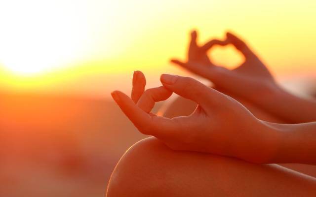 Person meditating outdoors at sunset with hands in meditation mudra, warm glow in background.