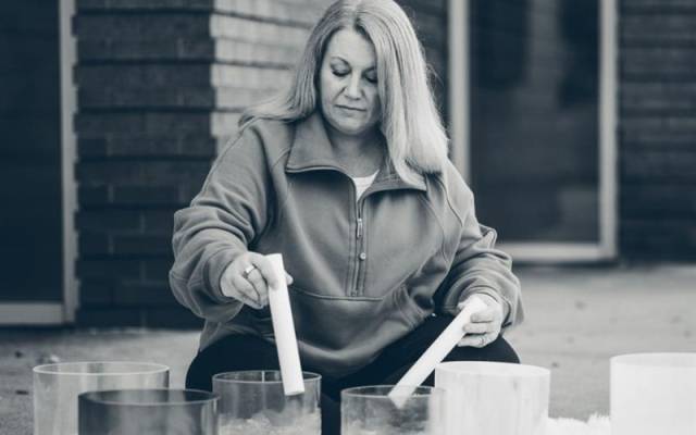 Person playing crystal singing bowls with mallets outside near brick wall and window.