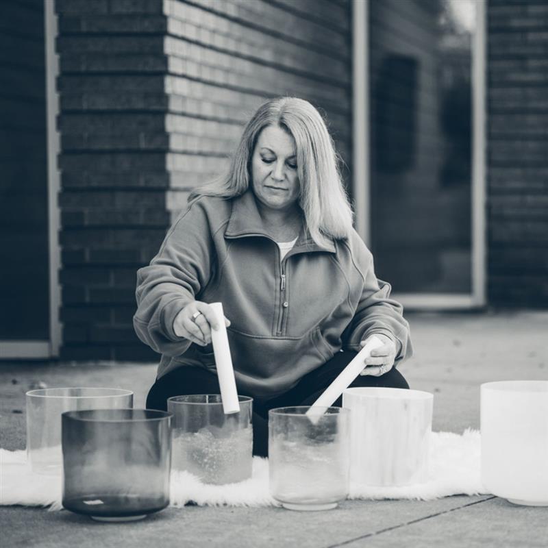 June _ Sound Bath Experience Person playing crystal singing bowls with mallets outside near brick wall and window.