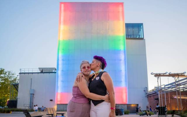 Couple embraces in front of a building with vertical rainbow lights, under a clear sky.