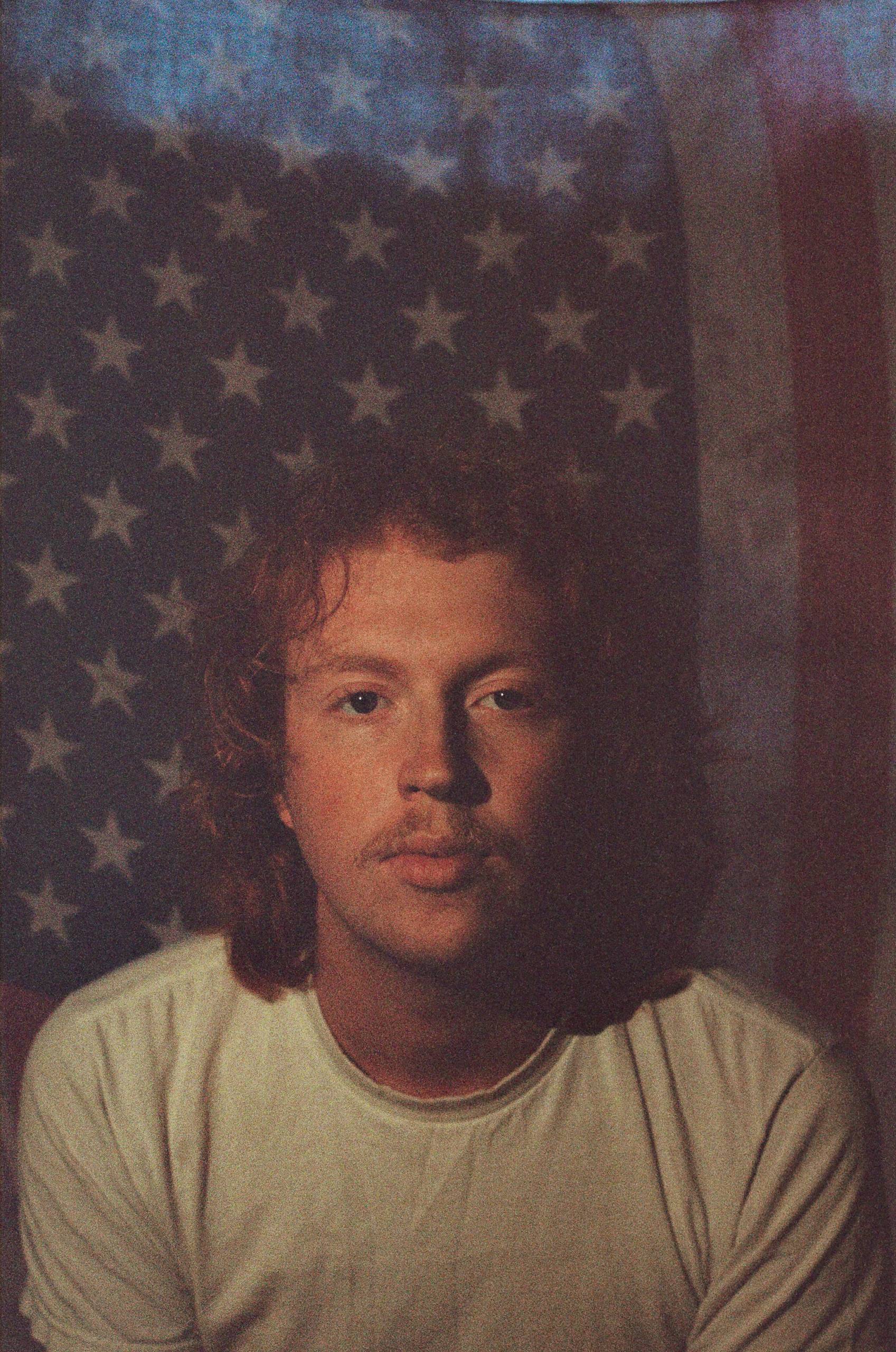 RYMAN Young man with curly hair and mustache wearing a light T-shirt in front of an American flag.