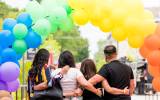 Four people embrace under a rainbow balloon arch with trees and urban background.