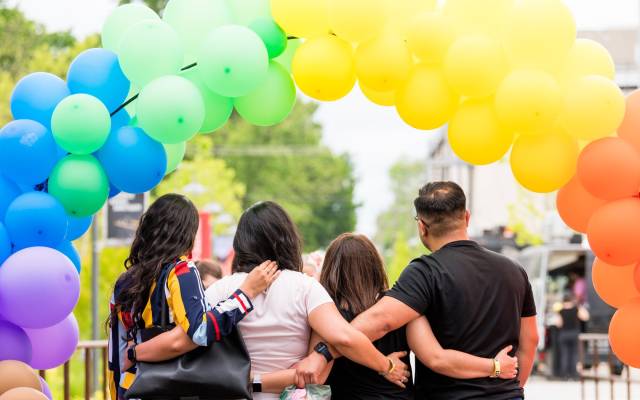 Four people embrace under a rainbow balloon arch with trees and urban background.
