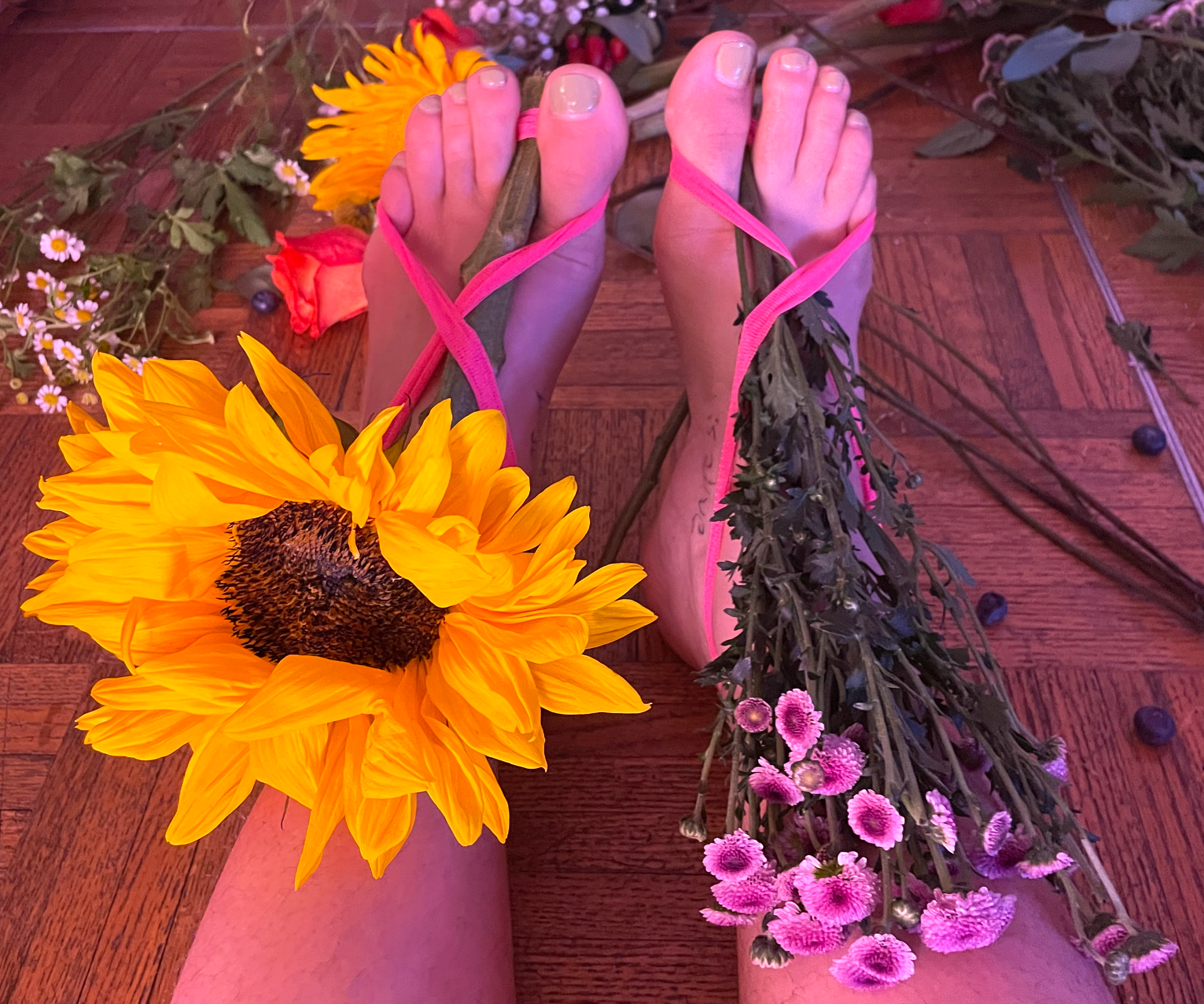 flower shoes-ashley Feet with pink nail polish and flowers, one with a large sunflower, on a wooden floor.