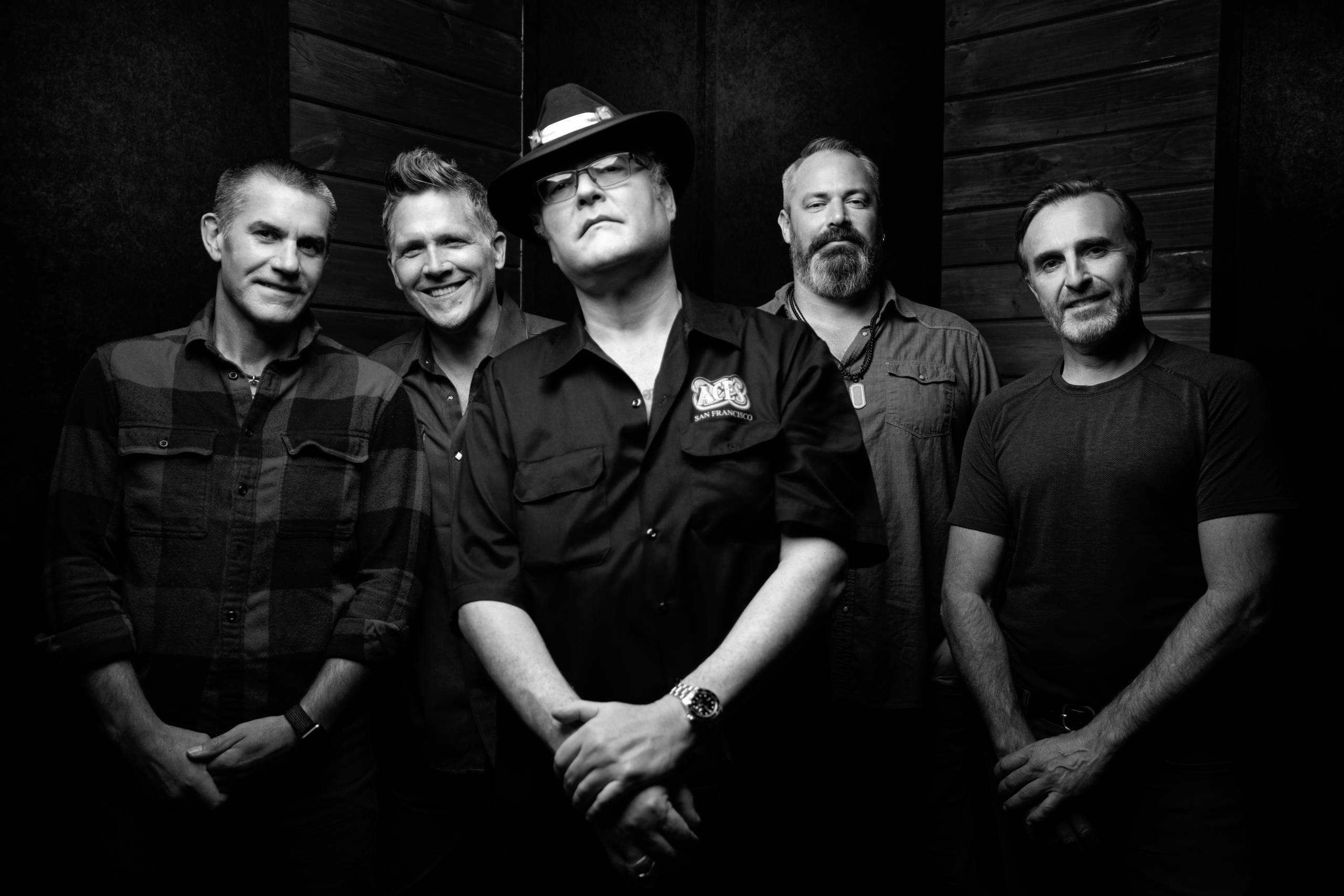 Blues Traveler Five men posed in a black and white photo with a wooden panel background.