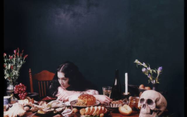 Person with head on table surrounded by food, skull, candle, and flowers in a still life setting.