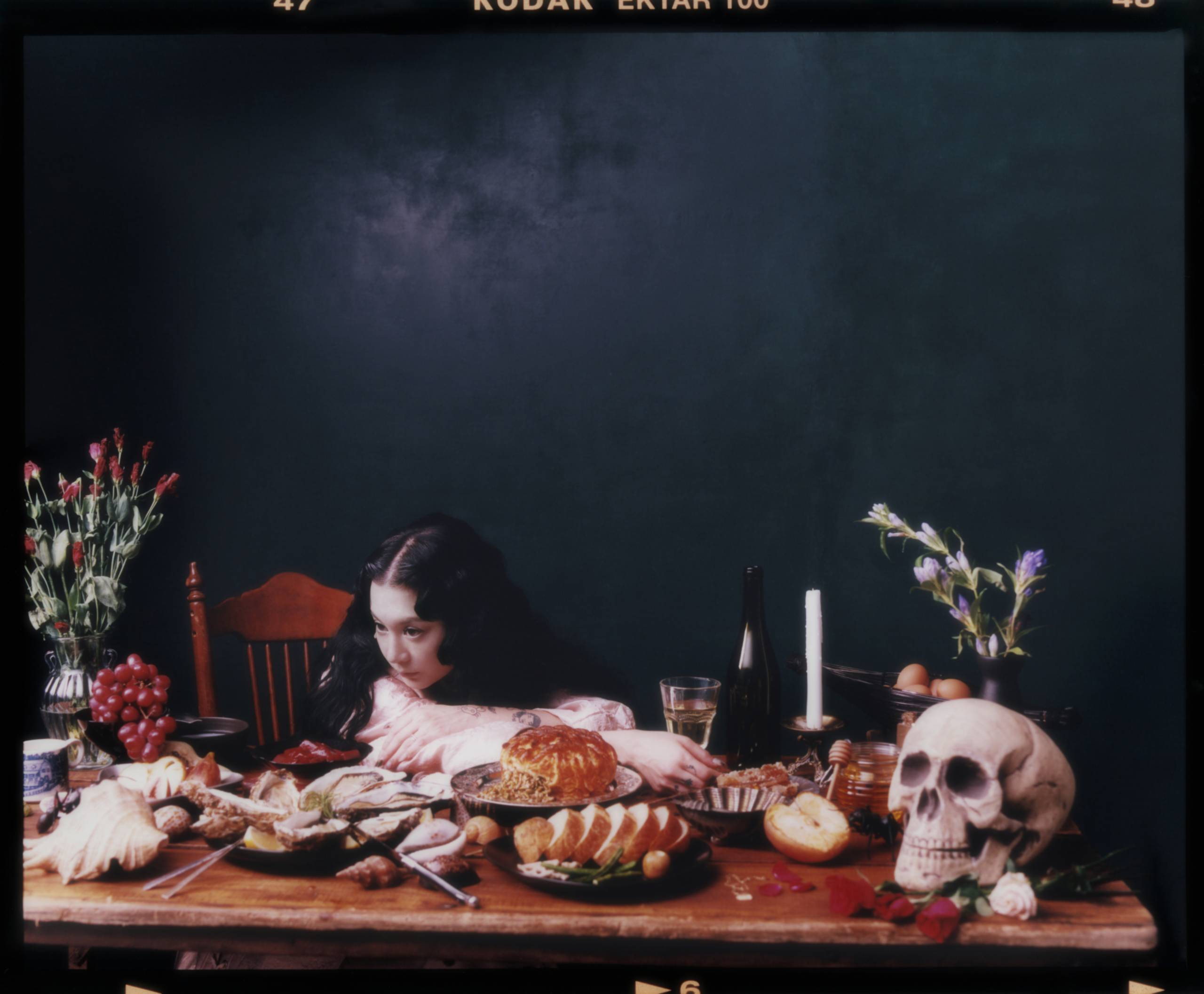 Japanese Breakfast Person with head on table surrounded by food, skull, candle, and flowers in a still life setting.