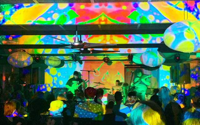 Band performing live with colorful psychedelic light projections on stage and ceiling.
