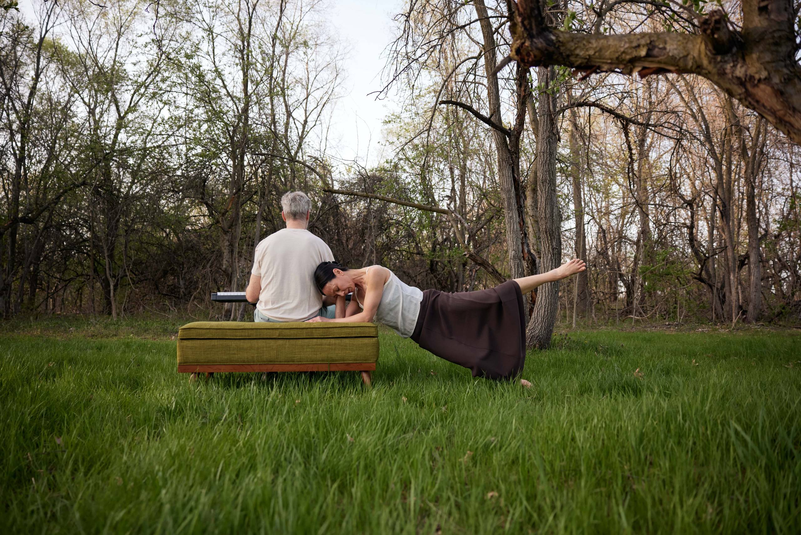 Robyn Mineko Williams - To Leave You Man in white shirt with back facing the camera sitting down on a bench outside playing an instrument. Next to the man is a woman in a brown skirt leaning against the bench with her fore arms and her legs fully extended outward.