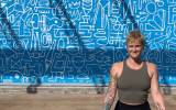 Person with tattooed arms in a meditative pose in front of a blue wall with white abstract drawings.