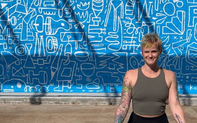 Person with tattooed arms in a meditative pose in front of a blue wall with white abstract drawings.
