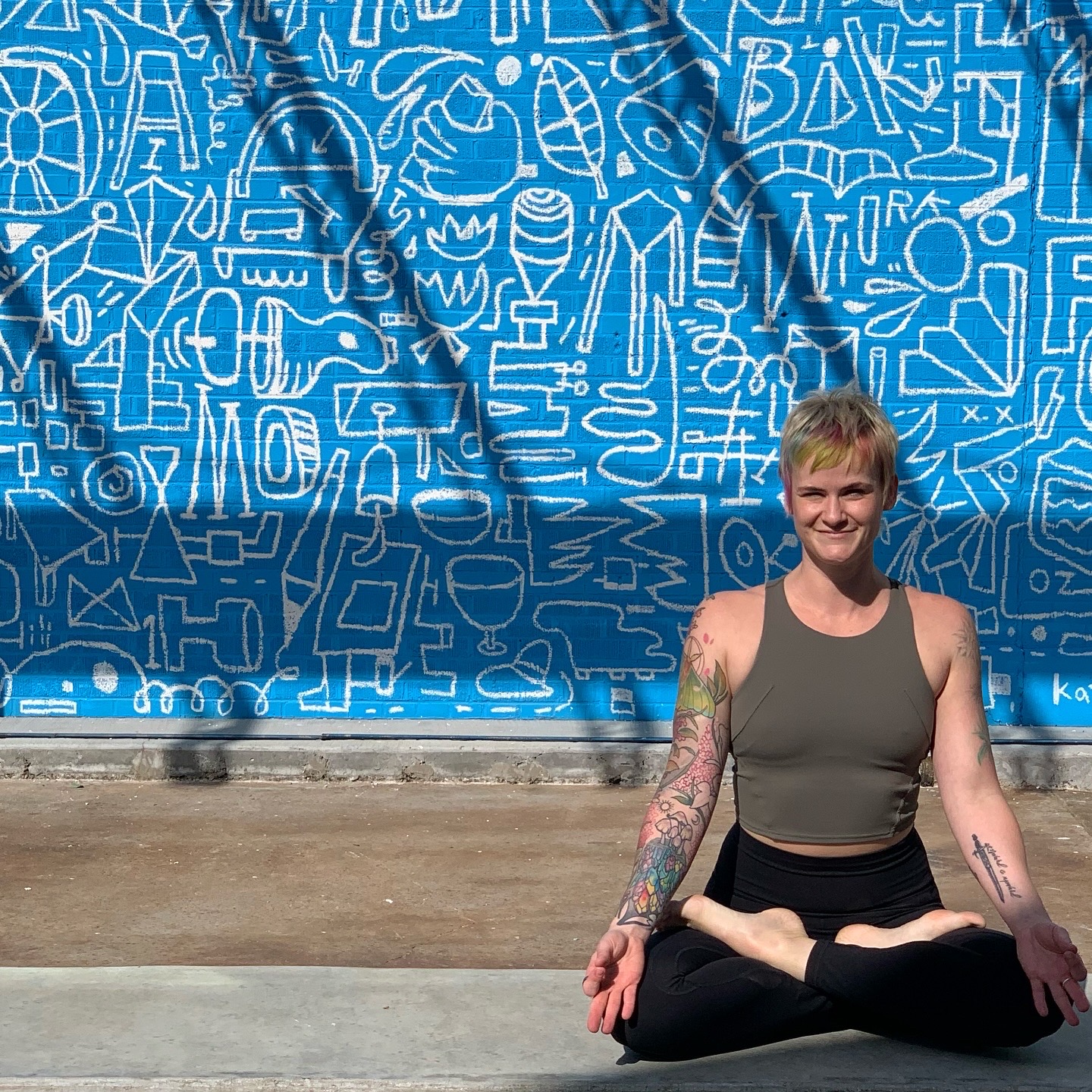 August Sun Reset 2025+ Person with tattooed arms in a meditative pose in front of a blue wall with white abstract drawings.