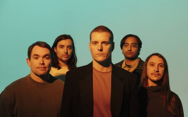 Five men posing closely together against a plain light blue background with warm lighting.