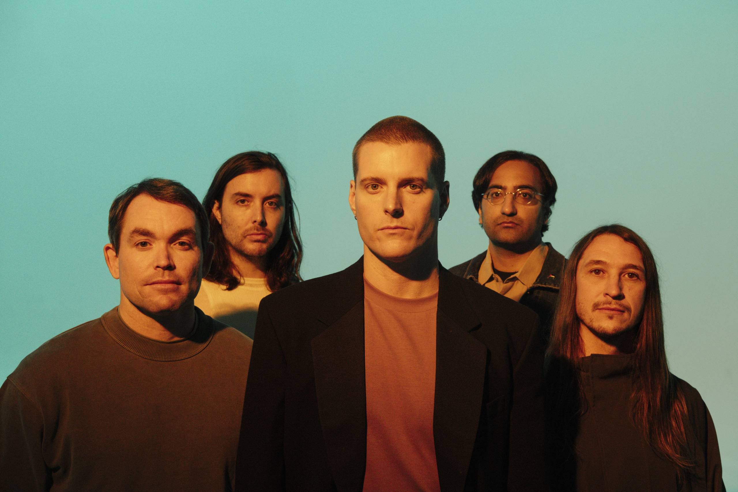 Deafheaven Five men posing closely together against a plain light blue background with warm lighting.