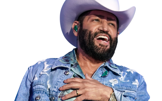 Man on stage singing in cowboy hat, denim jacket, patterned shirt, jeans, holding microphone.