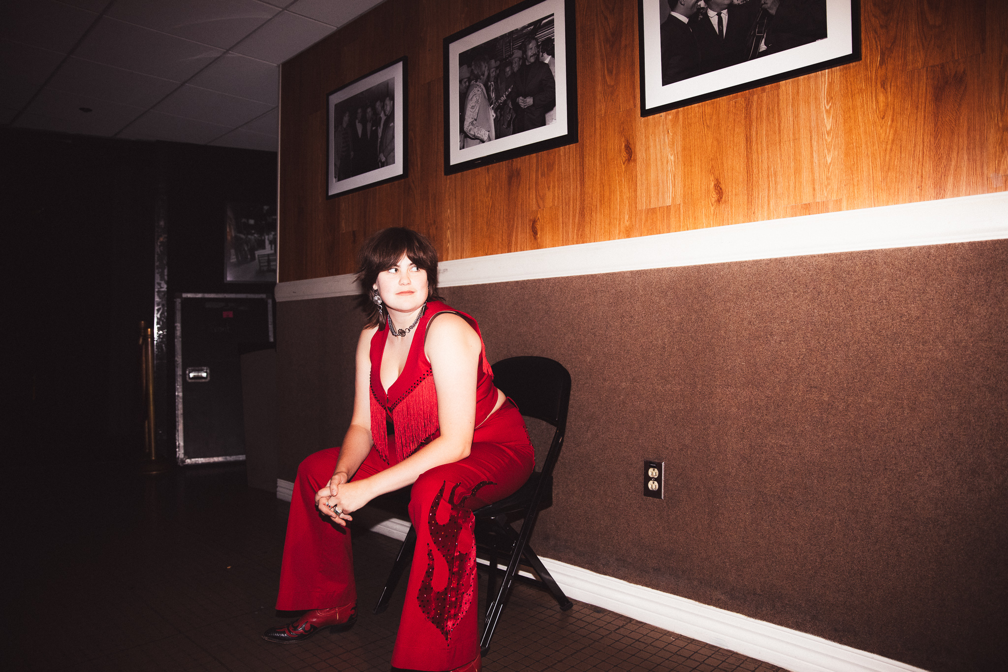 Person in red outfit with flame designs sitting on black chair against wood-paneled wall with photos.