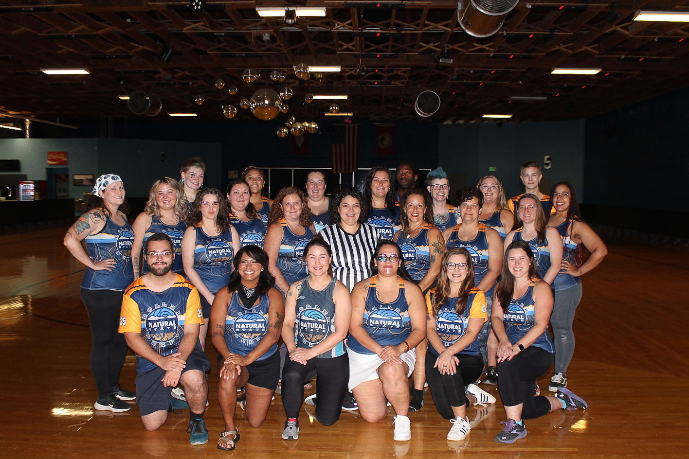 Natural State Roller Derby Group of people in blue and orange sports jerseys posing on an indoor wooden floor under disco balls.