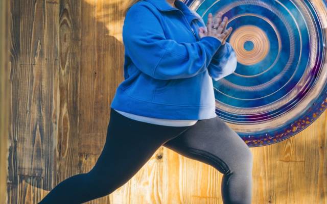 Woman in blue sweatshirt and black leggings doing yoga pose near a large gong on wooden floor.