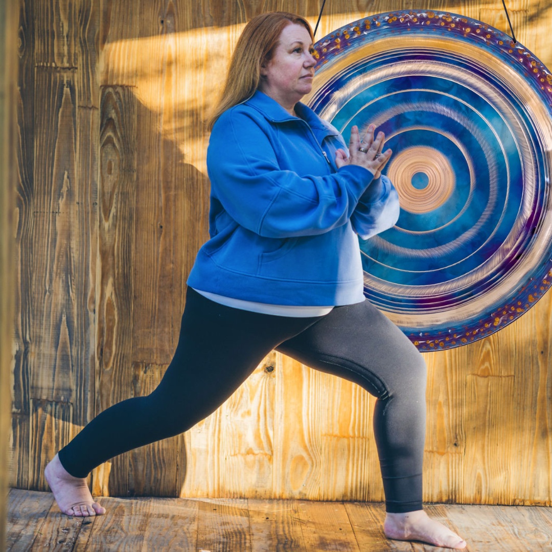 Woman in blue sweatshirt and black leggings doing yoga pose near a large gong on wooden floor.