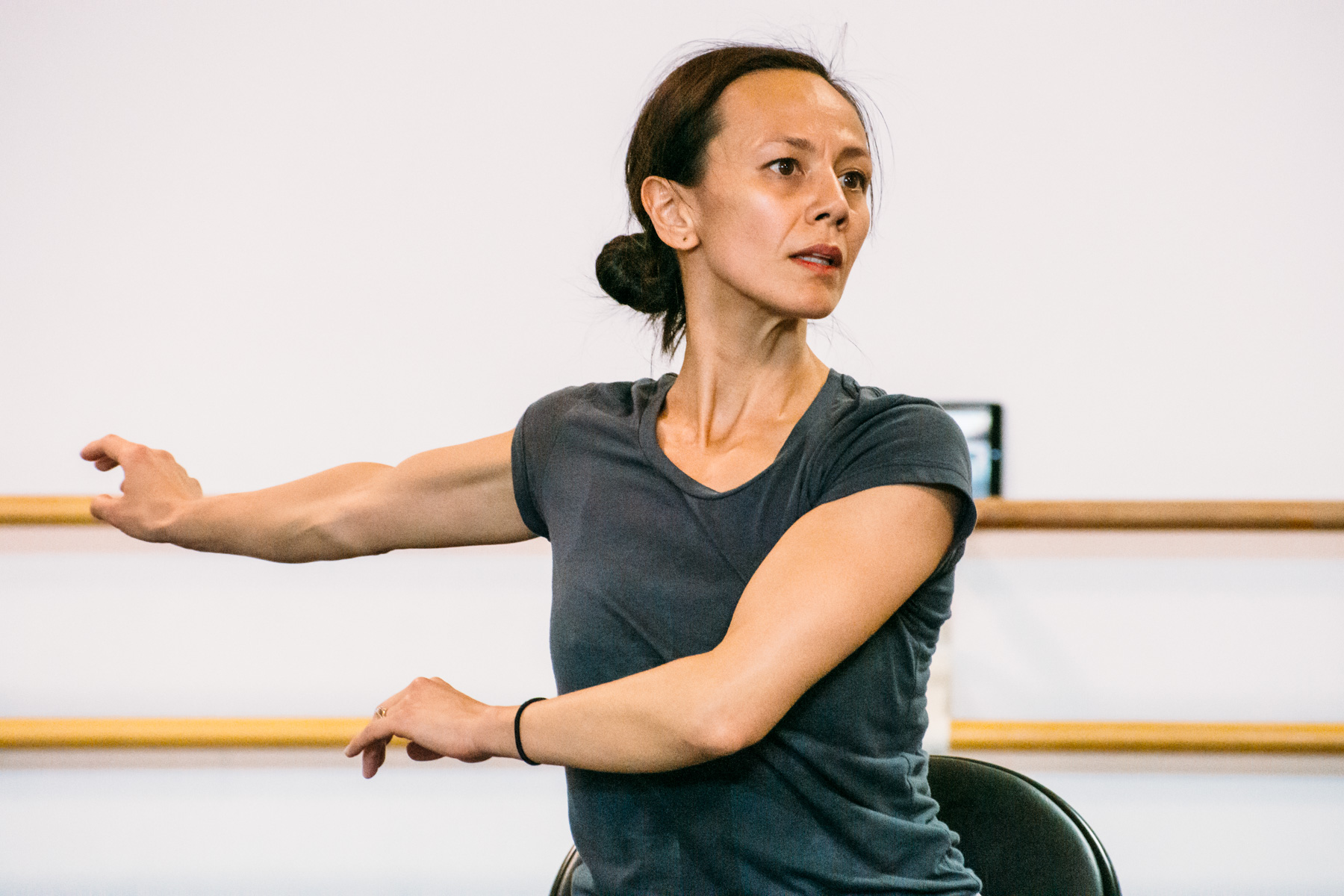 Robyn Mineko Williams Person with dark hair in gray shirt seated, arm extended in bright room with wooden railings.