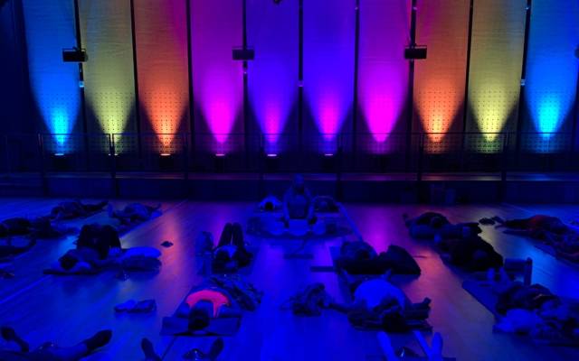 People lying on mats in a dim room with colorful gradient lights on the wall during a class.