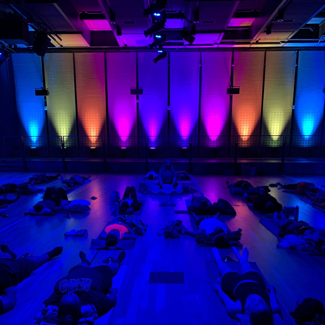 People lying on mats in a dim room with colorful gradient lights on the wall during a class.