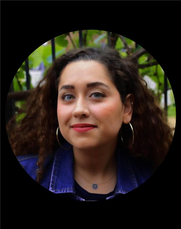 ela headshot Woman with curly brown hair wearing hoop earrings and a denim jacket outdoors with green foliage background