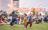 Children play on grassy area at an outdoor event, with tents and neon sign in background.