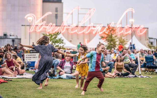 Children play on grassy area at an outdoor event, with tents and neon sign in background.