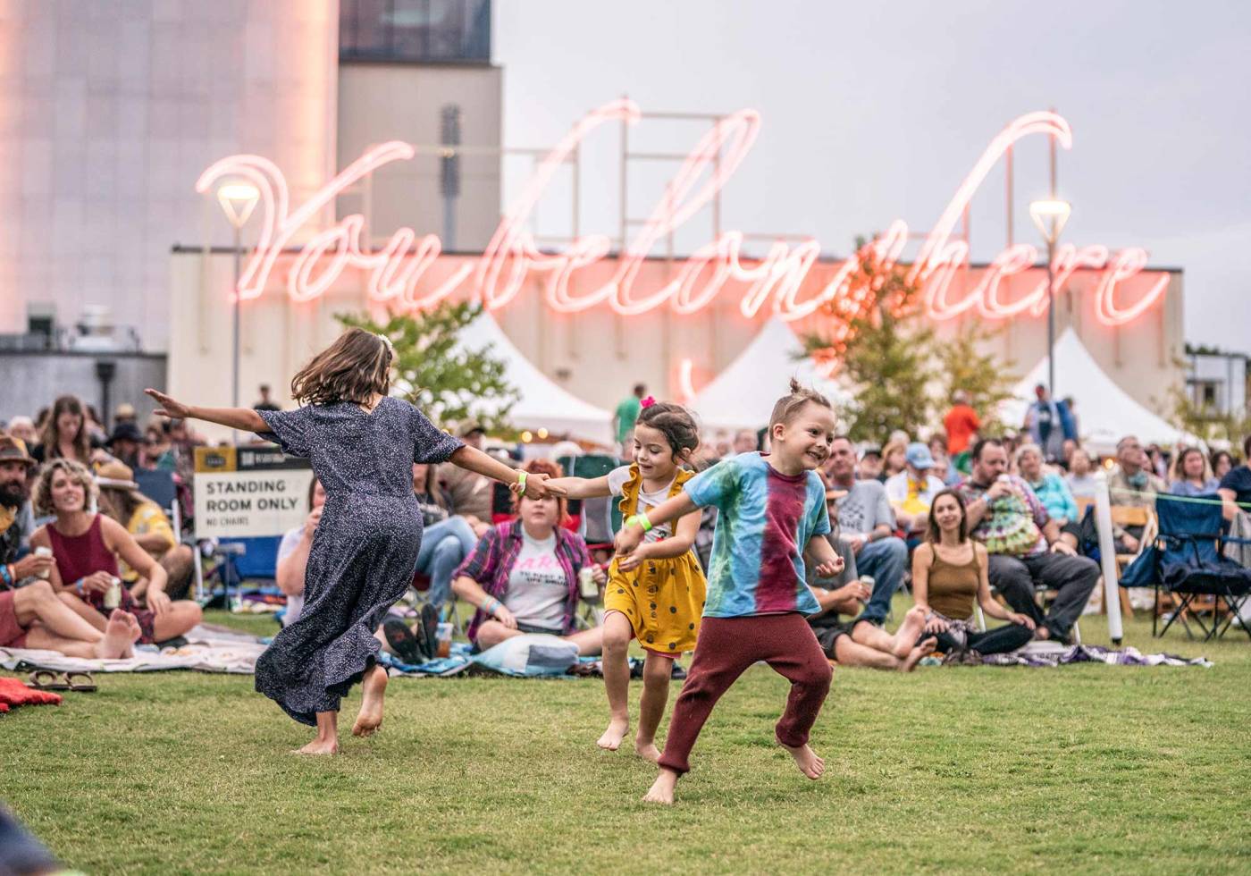 Children play on grassy area at an outdoor event, with tents and neon sign in background.