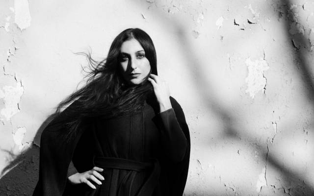 Black and white photo of a woman with long dark hair in a belted dark outfit by peeling wall.