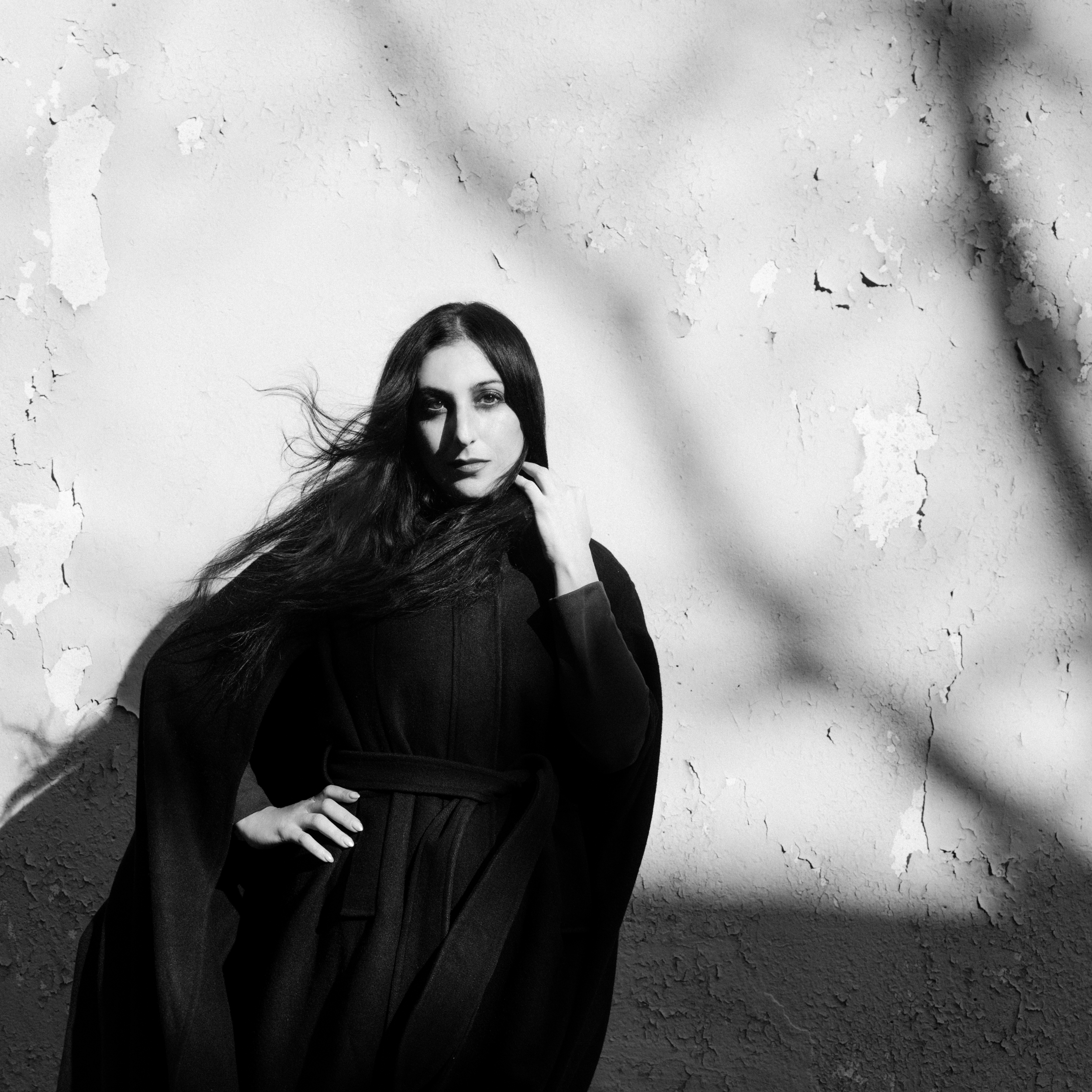 Black and white photo of a woman with long dark hair in a belted dark outfit by peeling wall.