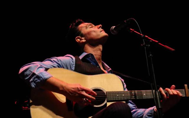 Man playing acoustic guitar and singing into microphone on a dark stage with red lighting.